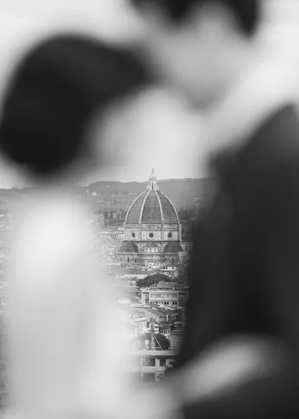 Couple portrait in black and white, the bride in a bridal gown, framed by a city skyline with a cathedral dome and distant hills beneath clouds.