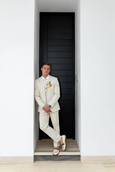 Groom portrait of a man in a cream suit with tie and yellow rose boutonniere, wearing a wristwatch, leaning by a black door in a white doorway