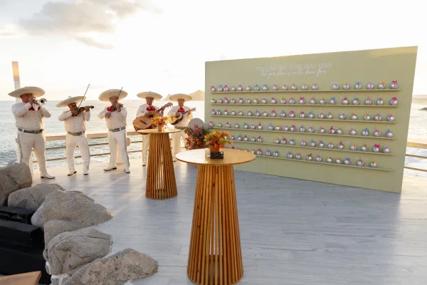 Wedding escort display with an escort card wall of ribbon place cards and favor bags on shelves, set on an oceanfront deck at sunset