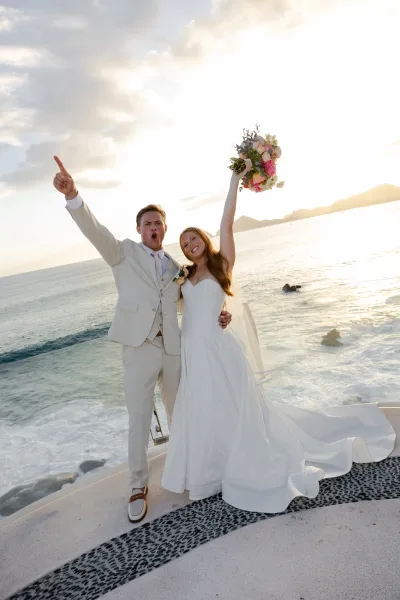 Couple portrait of bride and groom at a beach wedding couple cheering, bride lifting bouquet on an oceanfront terrace at sunset