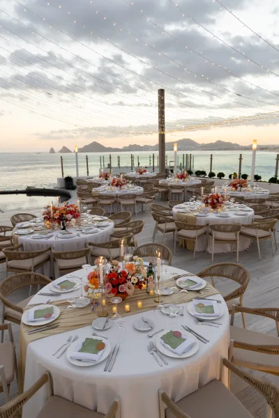 Reception tablescape for an outdoor wedding reception with round white-linen tables, floral centerpieces and candles on a sunset oceanfront terrace