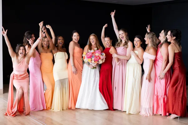 Bridesmaid group photo with bride with bridesmaids smiling in mismatched pink and coral dresses, holding a colorful bridal bouquet against a black wall