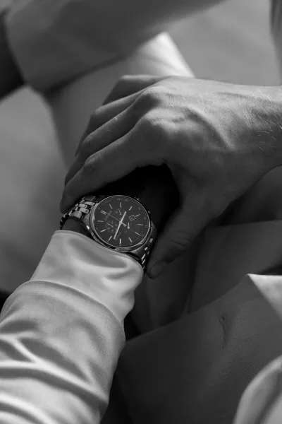 Groom watch detail with silver metal wristwatch peeking from a dress shirt cuff and suit sleeve, groom suit pants in soft blur