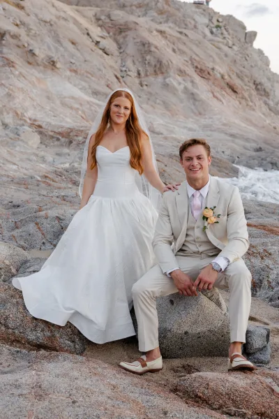 Couple portrait on a rocky beach wedding, bride in strapless gown and veil standing by seated groom in beige suit with boutonniere, ocean waves behind