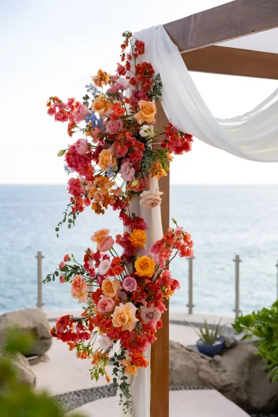 Wedding ceremony arch with white draping fabric and rose florals, set on a stone patio overlooking the ocean and sky backdrop