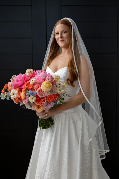 Bridal portrait of a bride holding bouquet with a long veil, strapless textured gown, and necklace against a black paneled wall