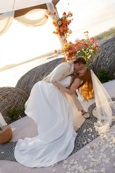 Wedding kiss portrait of bride and groom in a dip kiss wedding photo beneath a draped floral arch by the ocean at sunset light