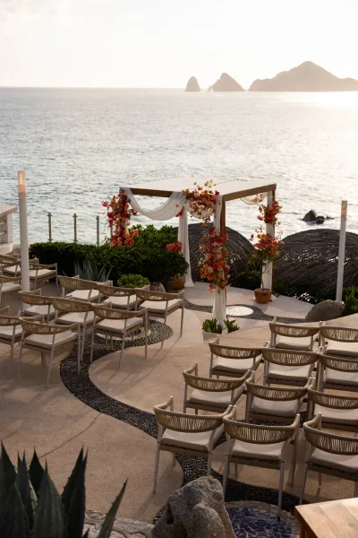 Ceremony setup for an oceanfront wedding ceremony with a wood wedding arch and white drape on a terrace overlooking rocky coastline at sunset