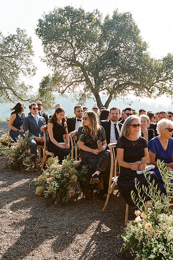 Wedding guests in formal attire seated in wood chairs, some wearing sunglasses, beside aisle flowers and greenery with mountain backdrop
