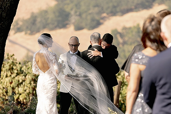 Ceremony moment as groom hugs his father beside the bride at the altar, veil flowing, with officiant and guests in a vineyard hillside setting