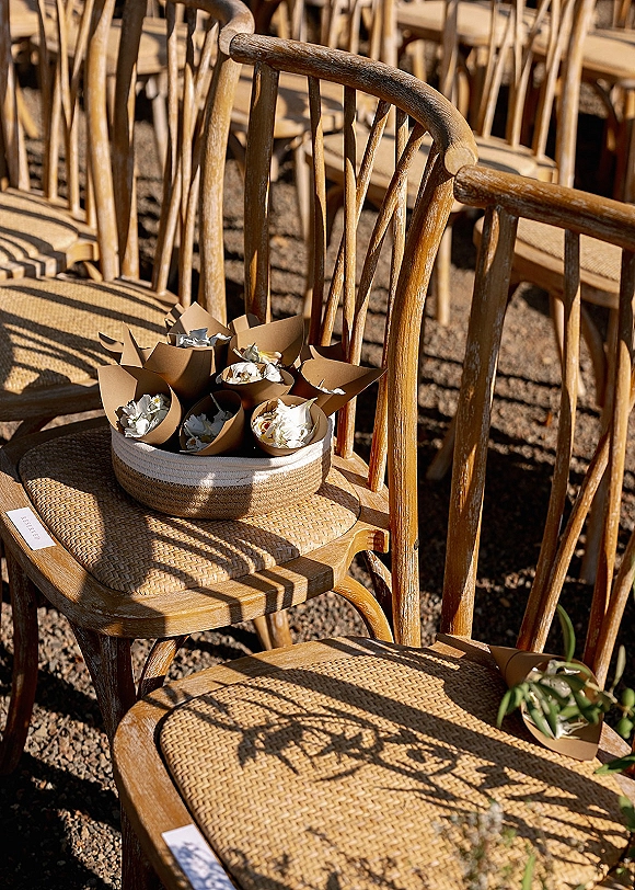 Ceremony seating decor with wedding aisle chairs featuring cane seats, reserved cards, petal cones and a basket on gravel in sunlit rows