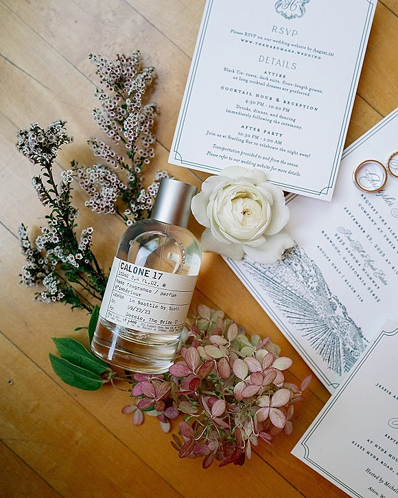 Invitation suite wedding invitation flat lay with RSVP and map insert, rings, perfume bottle, and white rose on a wood floor background