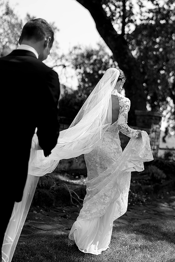 Wedding processional with bride walking away in a lace long-sleeve gown and cathedral veil blowing, groom in tuxedo on a garden path