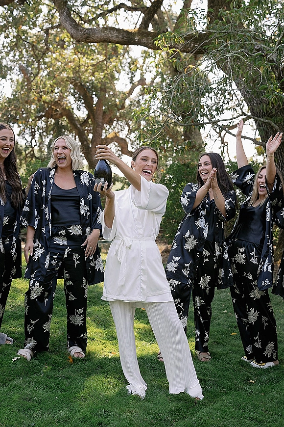 Bride with bridesmaids in matching bridesmaid pajamas, laughing on a grassy lawn under tree canopy as champagne bottle pops in daylight