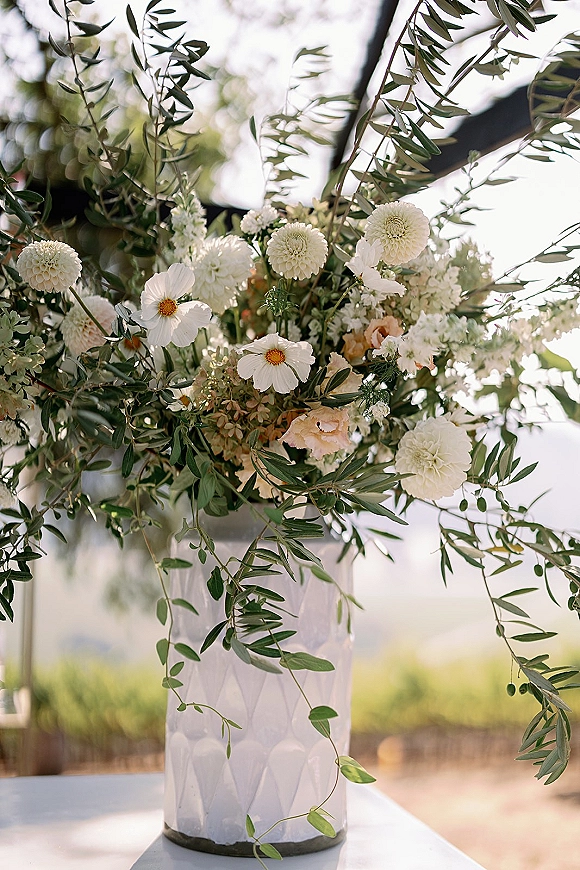 Wedding centerpiece with tall white dahlias and blush roses in a white ceramic vase, accented by olive branches on a sunlit outdoor table