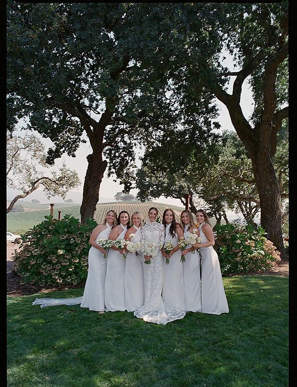 Bridesmaids portrait with bride in lace gown and veil, all-white dresses and white rose, calla lily, and greenery bouquets in a vineyard lawn setting