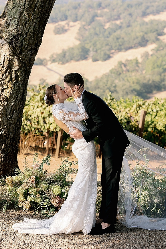 Wedding kiss portrait of bride and groom kissing under a tree, her cathedral veil and lace sleeves flowing with vineyard hills behind