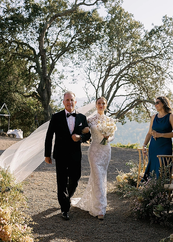 Wedding processional as bride walking down aisle with her father, holding white bouquet and veil train, past aisle florals on a gravel path outdoors