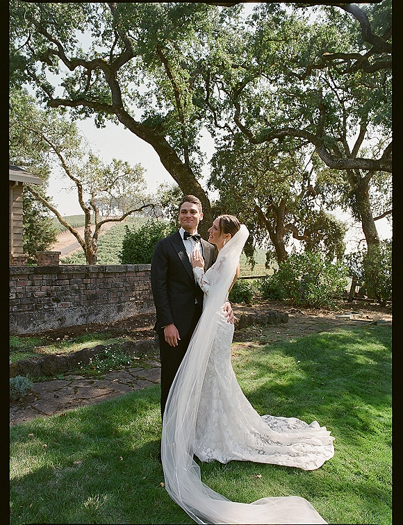 Couple portrait of bride in lace gown with long veil and train, touching groom’s tuxedo lapel on a garden lawn by oak trees