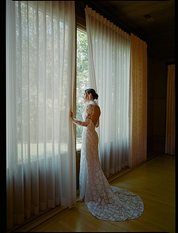 Bridal portrait of a bride by window, looking out in a long sleeve lace open-back gown with train, holding sheer curtains in natural light