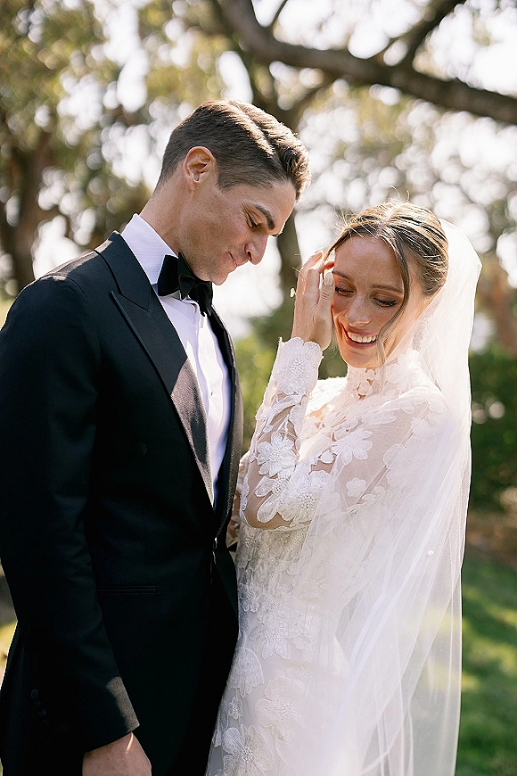 Couple portrait of bride in long sleeve lace dress and veil with groom in tuxedo and bow tie, standing in sunlit trees and greenery