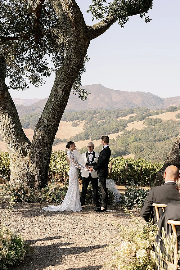 Wedding vows at an outdoor wedding ceremony as bride in lace long sleeves and groom in tux hold hands under an oak tree in a vineyard