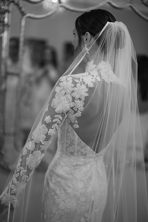 Bridal portrait of a bride in a backless wedding dress with cathedral veil and lace sleeves, looking away by an ornate mirror indoors