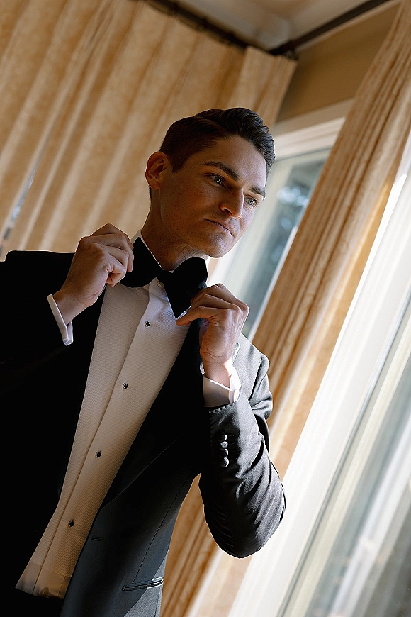 Groom portrait in a black tuxedo adjusting his bow tie, white waistcoat and cufflinks visible, standing by window light with sheer curtains