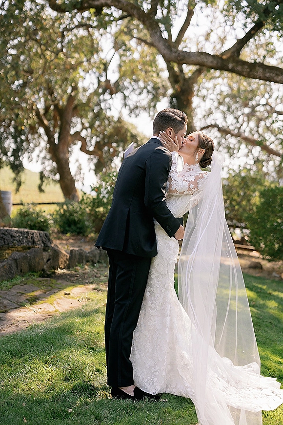 Wedding kiss portrait of bride and groom kissing, her long veil blowing in sunlight beneath oak trees on a grassy lawn with rocks