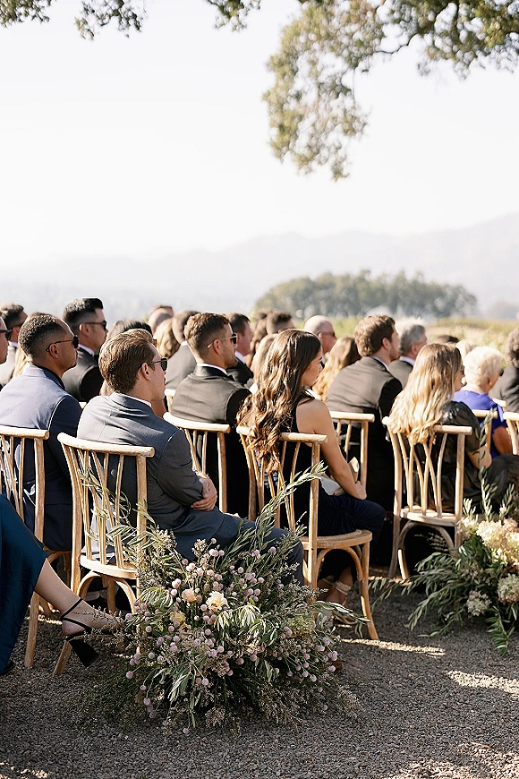 Ceremony guests seated in rows on wood chiavari chairs beside aisle florals and greenery garlands, facing a mountain landscape under bright sky