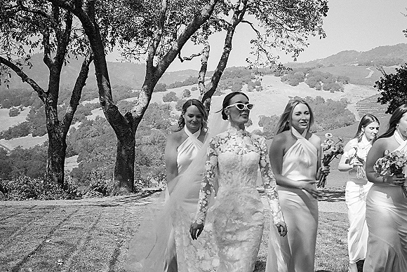 Bridal party portrait of bride with bridesmaids walking downhill in oak shade, bride in long-sleeve lace gown and cat-eye sunglasses, holding bouquets