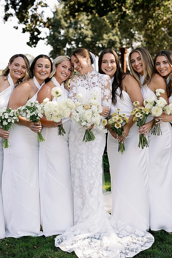 Bride with bridesmaids in mismatched white dresses holding white rose and greenery bouquets, with lace gown and cathedral veil in a sunny garden
