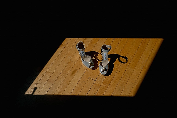 Bridal shoes in a wedding shoe flatlay, white open-toe ankle strap heels with pearl embellishments on a sunlit wood floor shadow