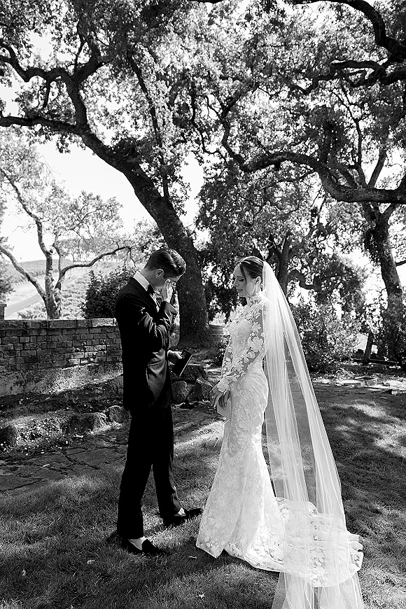 First look moment as groom crying first look, wiping tears while bride in lace gown and long veil stands by stone wall under oak trees