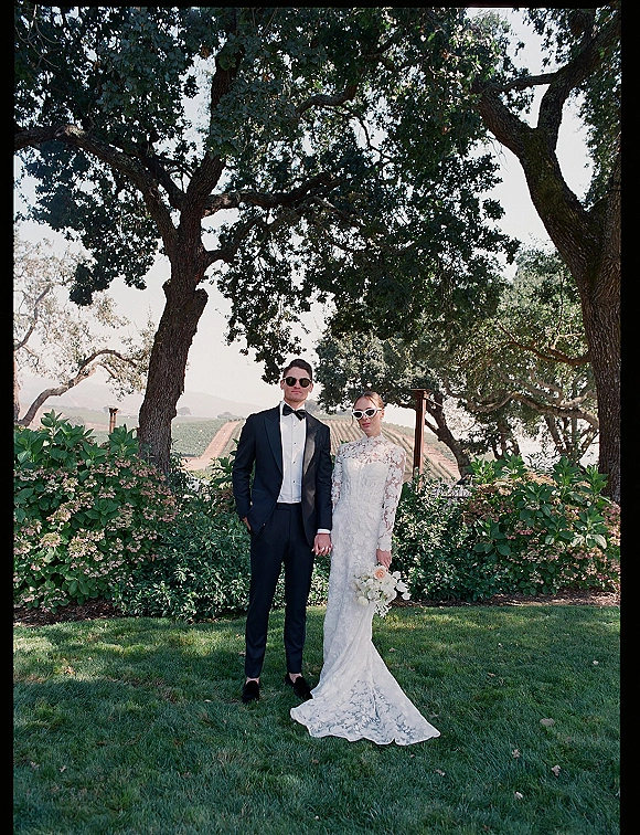 Couple portrait of bride and groom holding hands in sunglasses, she in long sleeve lace dress with white bouquet in vineyard garden setting