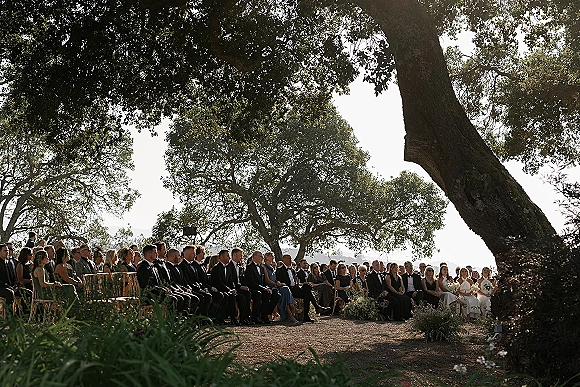 Ceremony guests at an outdoor wedding ceremony seated in wood chairs, black tuxedos and bridesmaids with bouquets under an oak tree canopy