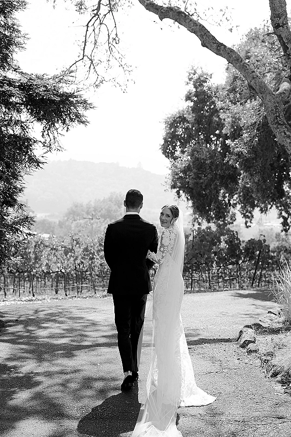 Couple portrait in a black and white wedding photo, bride in lace sleeves and veil walking with groom along a vineyard path with hills