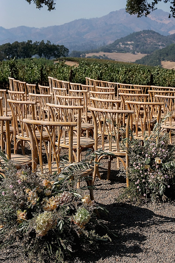Ceremony seating with wood chairs lined along a gravel aisle, greenery garland and floral arrangements set between vineyard rows and mountains