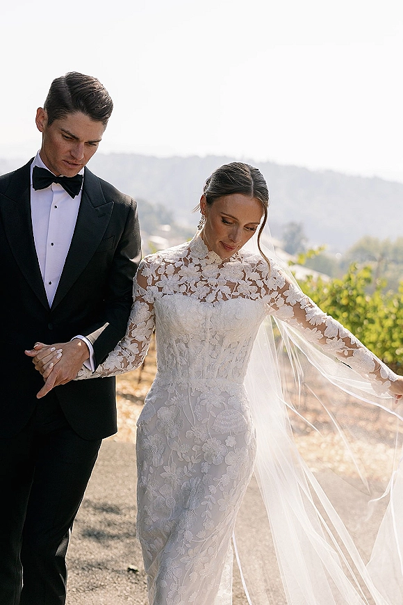 Couple portrait of bride holding groom’s arm as they walk a vineyard path, her long lace veil trailing, mountains behind them