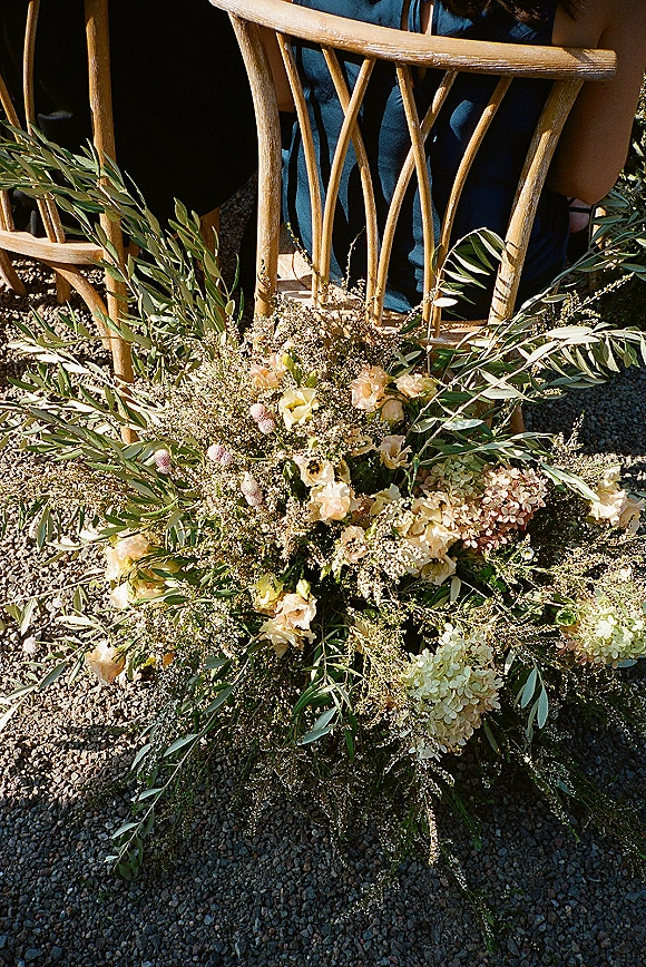 Aisle florals in a ground floral arrangement of hydrangea, greenery, and blush-white blooms beside a wooden chair on gravel with guests seated
