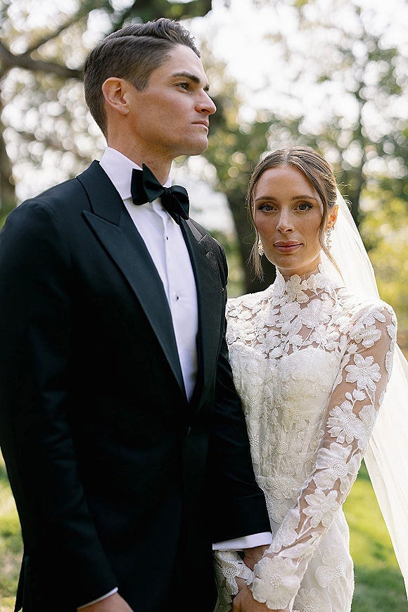 Couple portrait of bride in a high-neck long-sleeve lace gown and veil with groom in black tuxedo, standing in sunlit greenery