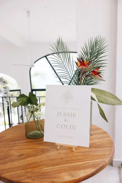 Welcome sign on a mini easel with tropical greenery and bird of paradise in a glass vase on a round wood table by an arched window