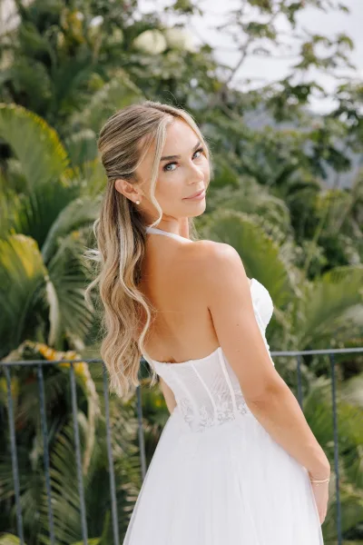 Bridal portrait of a bride looking over shoulder in a strapless lace corset gown on a terrace balcony with tropical greenery and palms behind.