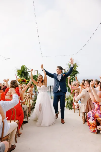 Wedding recessional as bride in strapless gown and groom in navy suit cheer hand in hand down a white runner on an ocean-view terrace