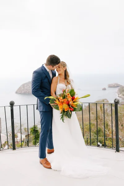 Couple portrait of bride and groom embracing with foreheads touching, bride holding bouquet by a railing above ocean and rocky islands