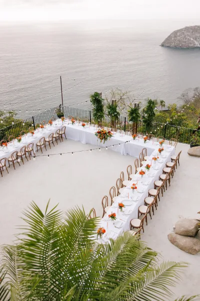 Reception tablescape with U shaped banquet table in white linens, bentwood chairs, orange florals, candles, and string lights on a terrace ocean view