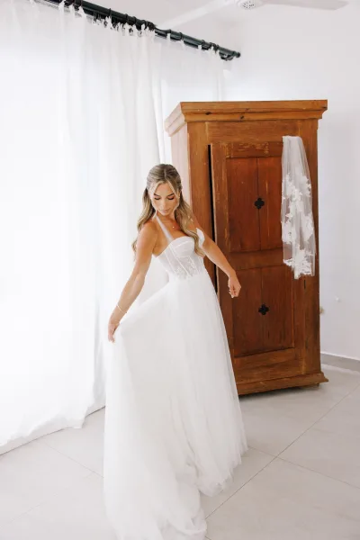 Bridal portrait of a bride in a wedding dress with veil, holding her tulle skirt by sheer curtains beside a wooden armoire indoors