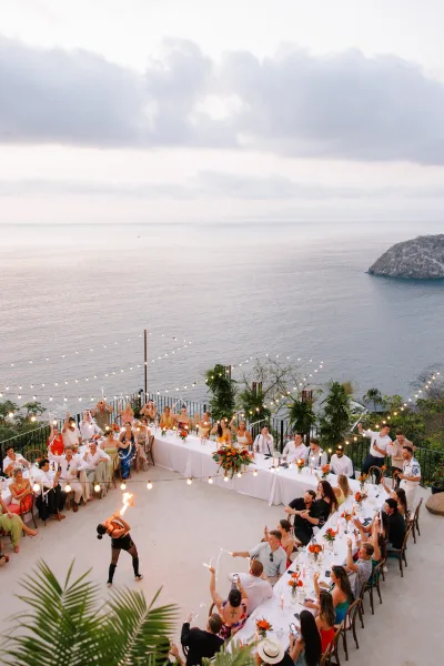 Reception entertainment as a wedding fire dancer performs with torches beside U-shaped banquet tables under string lights on an ocean-view terrace