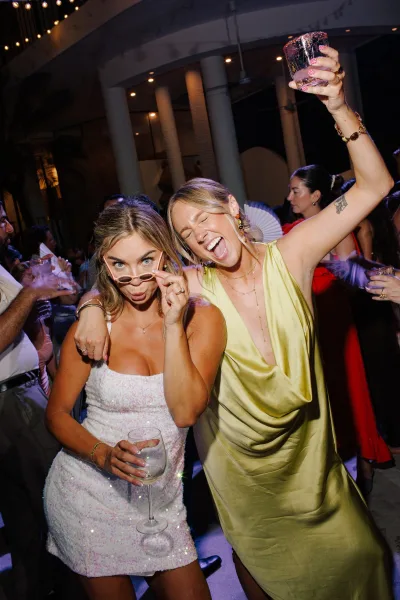 Wedding dance floor packed with wedding reception dancing guests holding a champagne coupe as string lights glow beside tall venue columns