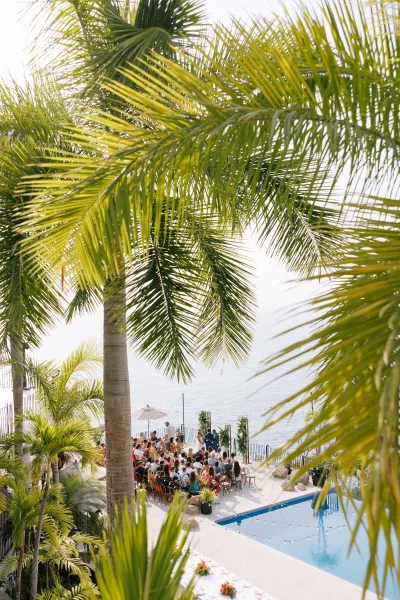 Outdoor wedding ceremony with destination wedding ceremony seating, white chairs and floral arrangements on a terrace by a pool with ocean view and palms